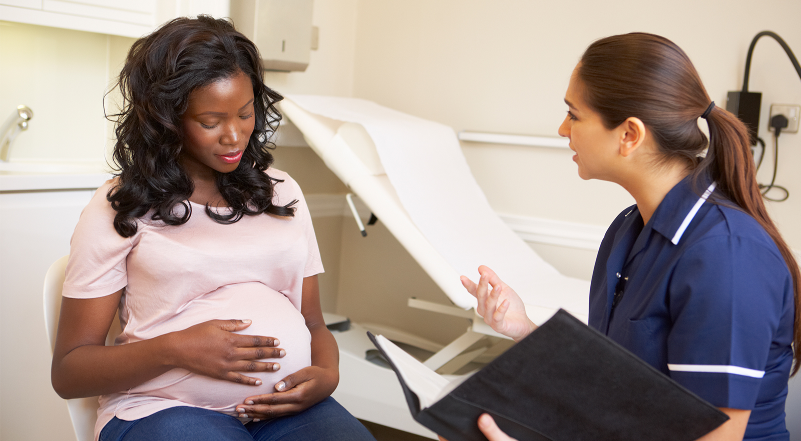 Image of a Pregnant Mother with her hands placed over the baby in her tummy in the doctors office talking to medical staff